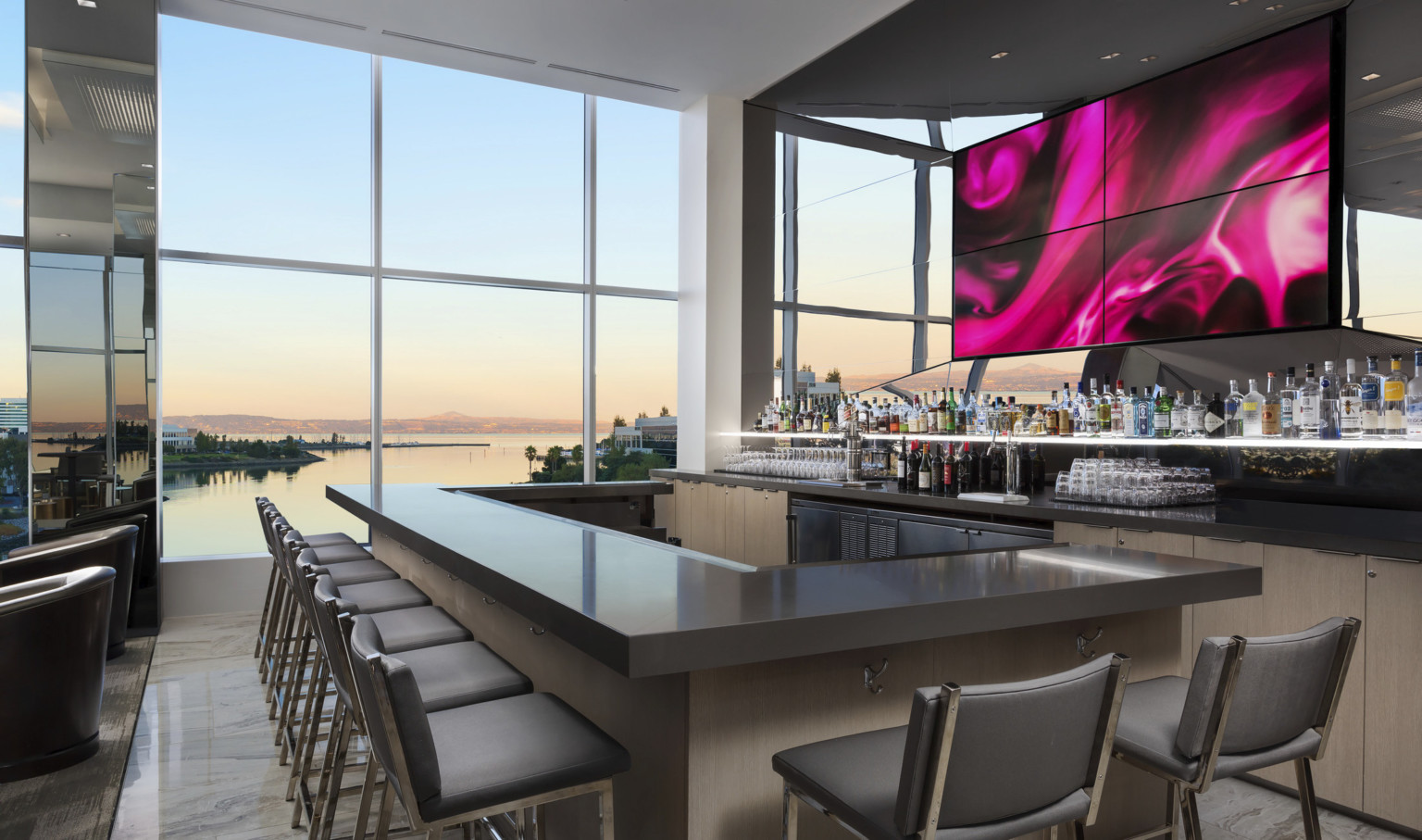 Wood bar with grey counter in room with floor to ceiling windows in front of mirror with central abstract swirling panels Wood bar with grey counter in room with floor to ceiling windows in front of mirror with central abstract swirling panels