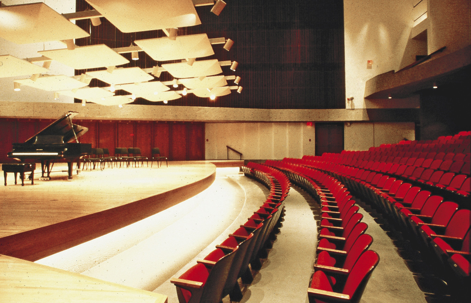 View inside UNO Performing Arts Complex theater. Rows of red seating face piano on stage under lights and acoustical panels