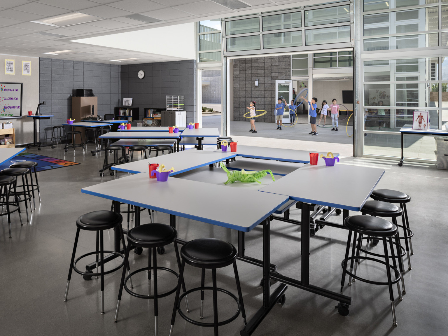 Classroom with flexible furniture, floor to ceiling windows, glass garage door raised to paved outdoor space between classes