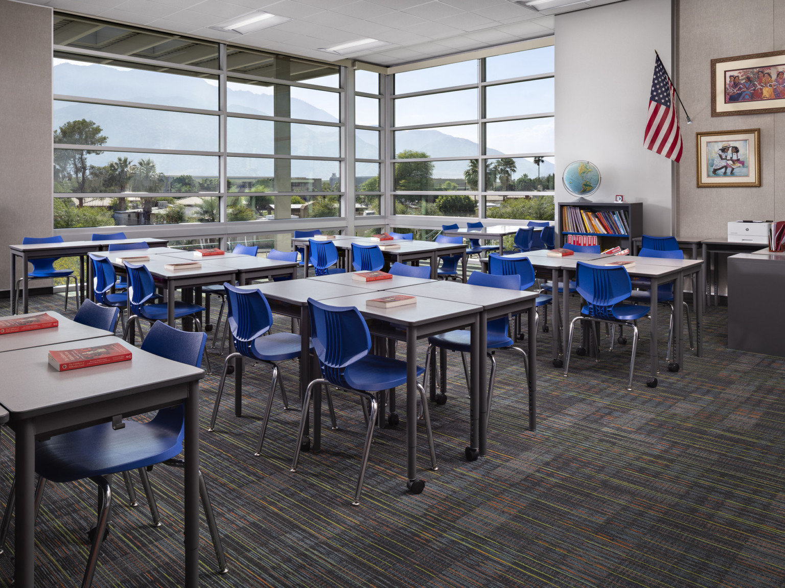 Classroom with flexible desks in quads with blue chairs. Floor to ceiling windows wrap around corner. Art on grey wall