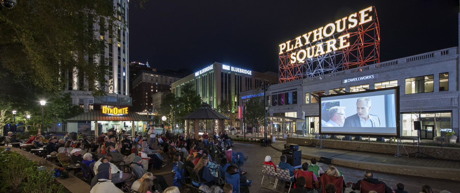 Audience sits on the street watching an outdoor movie on a screen. A Playhouse Square sign is illuminated in the background