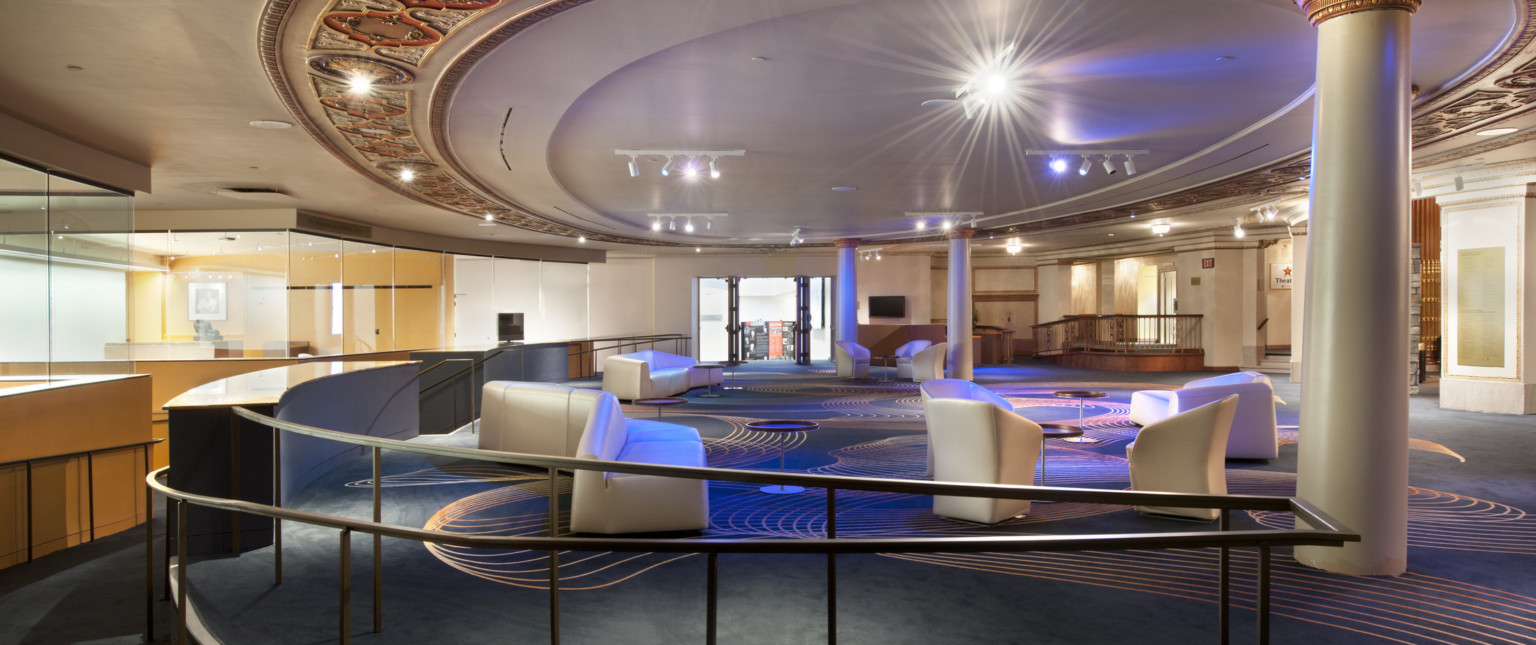 Lobby with dark colored carpet with gold organic design. White angled seats through out the room below a round ceiling detail