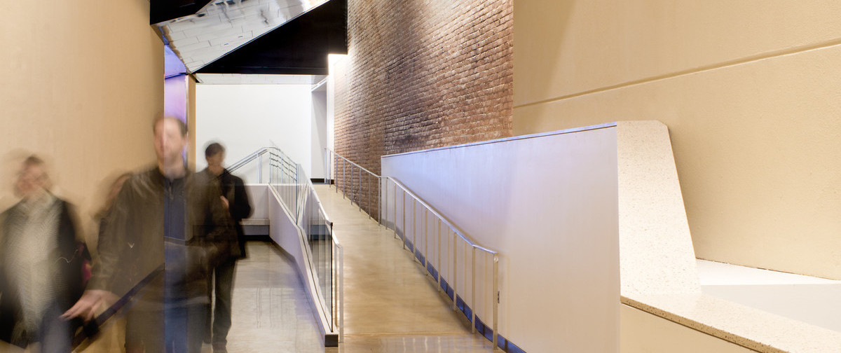 Hallway with ramp to the right, concrete yellow walls with exposed brick section reflected on metal ceiling section