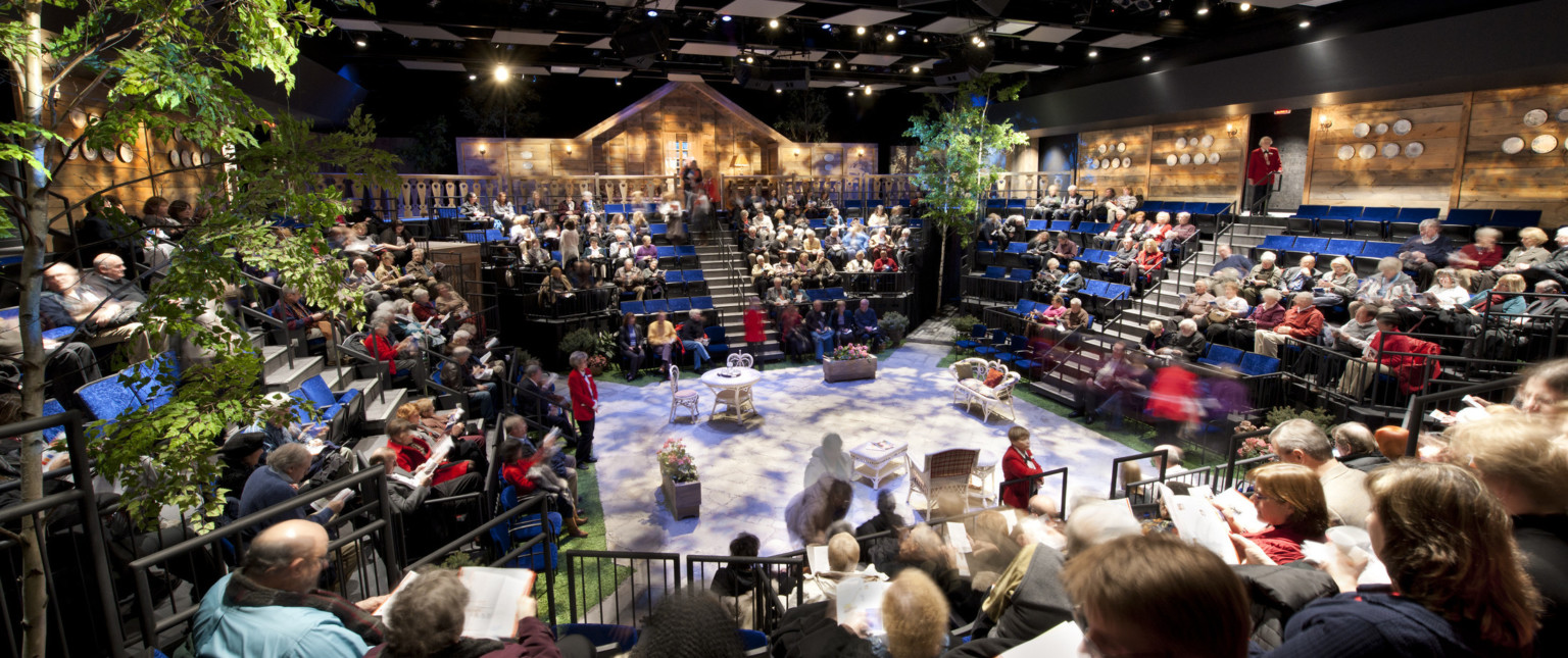 people seated at theater in the round with trees off a stage set with white table and chairs, textural lighting from above