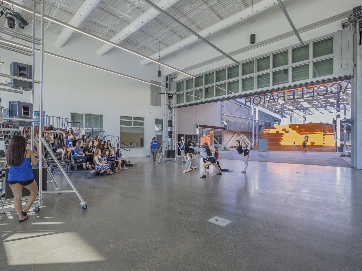 A white room, with the wall opened facing orange stairs, has portable bleachers and equipment