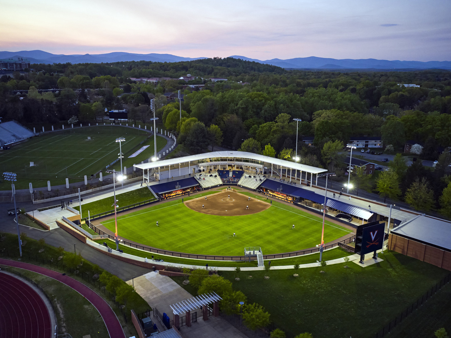 University of Virginia Softball stadium aerial view at lit up at dusk, surrounded by trees and view of Blue Ridge Mountains