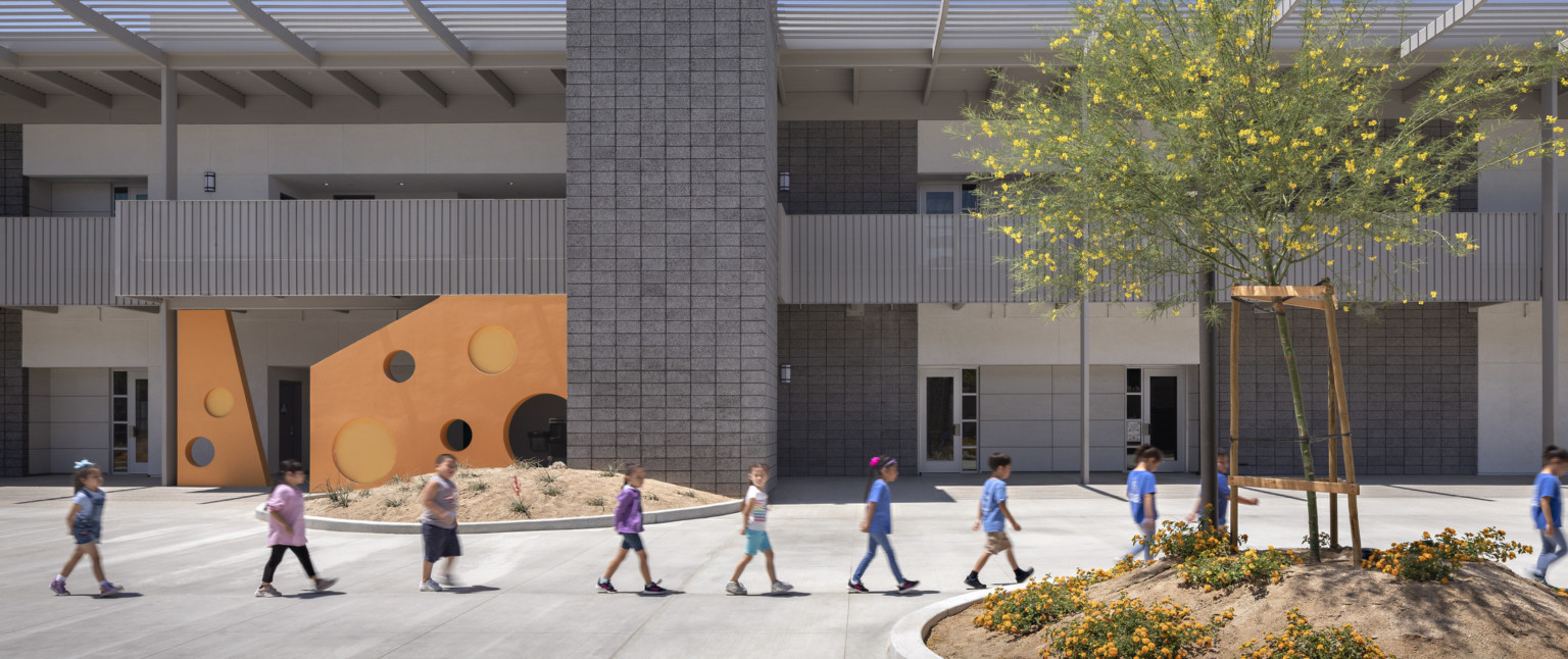 Children walk in a line through school courtyard in front of grey stone building with orange accent wall with round cut outs