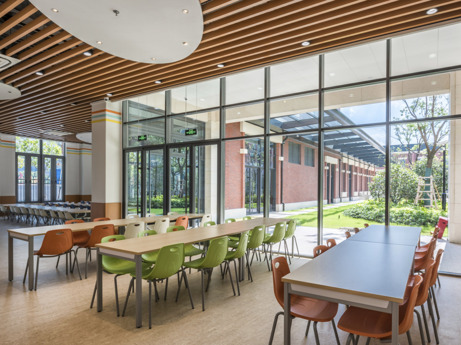 Dining area with long tables and colorful chairs looking out from floor to ceiling windows onto a courtyard with trees