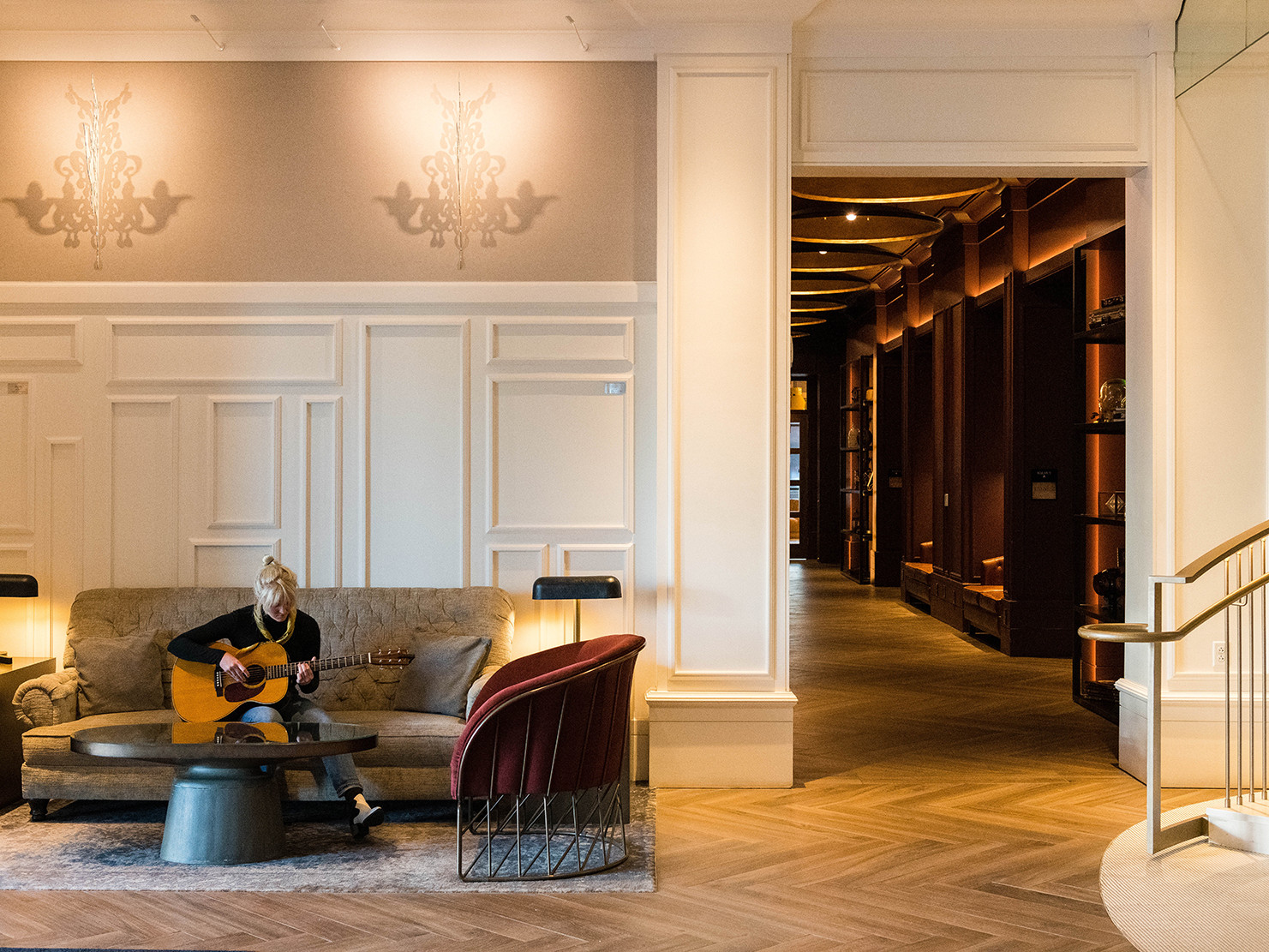 wood moldings in neutral tones with chandelier outlines on walls and herringbone floors. A woman plays guitar on gray sofa wood moldings in neutral tones with chandelier outlines on walls and herringbone floors. A woman plays guitar on gray sofa