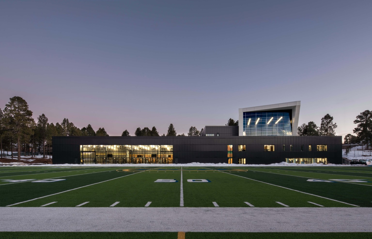 football field in front of a single story building with a multi story angular volume behind it against tree line sky at dusk