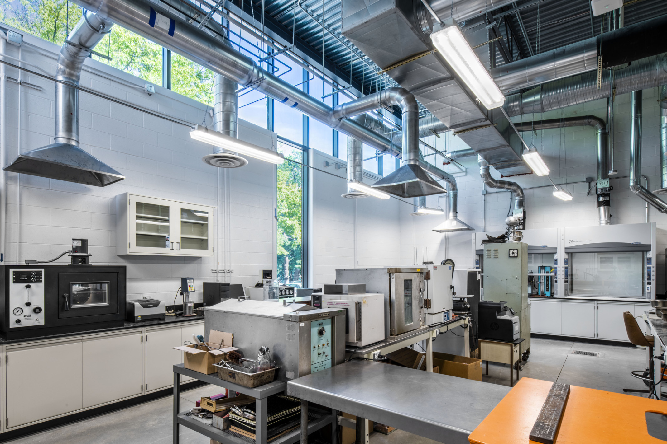 Double height white cement block lab with exposed ducts above lab equipment. View, left, through windows to green space