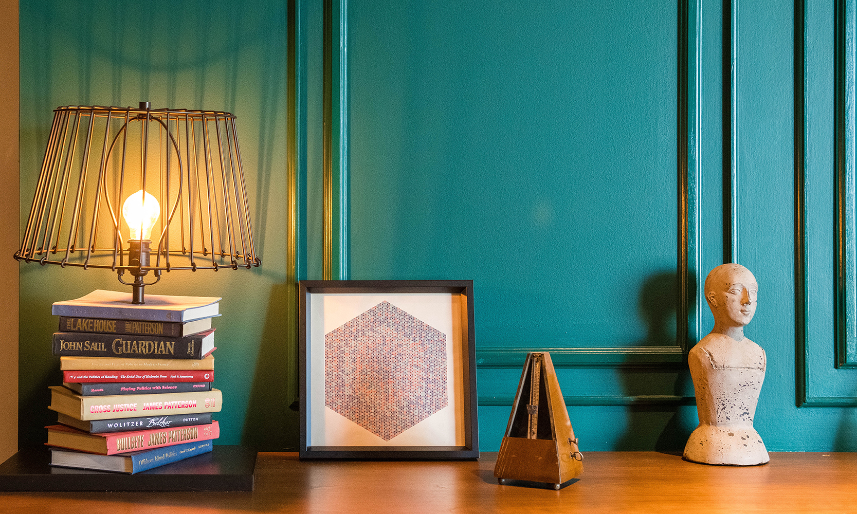 dark green wall with molding details behind a lamp made from stack of books, framed art print vintage metronome and cast bust