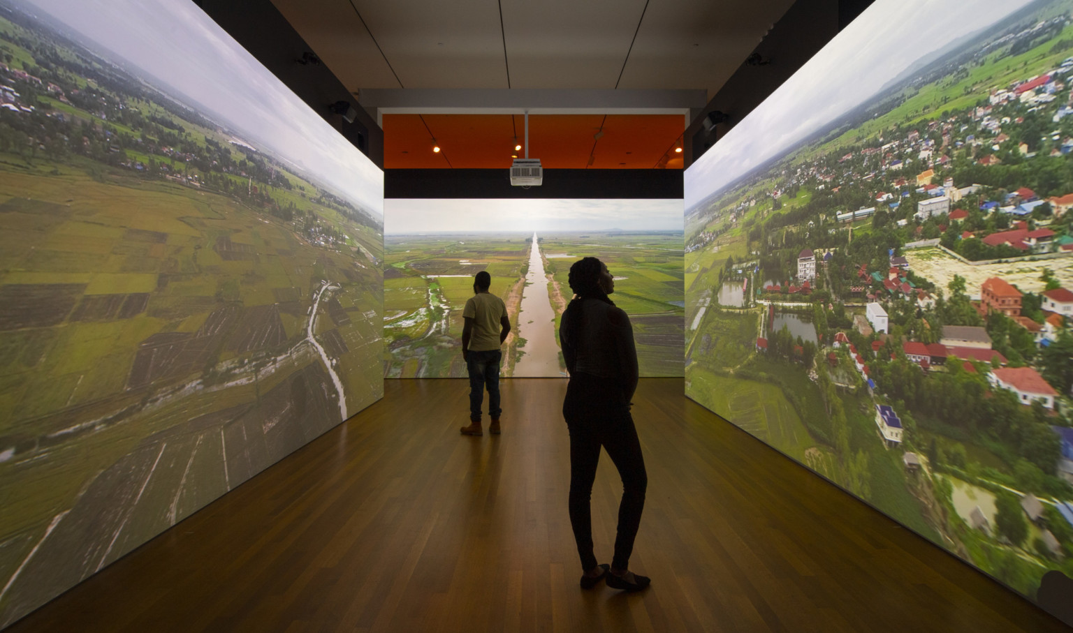 two darkened silhouettes stand in room with three walls with aerial images of farm lands, houses, and a river projected two darkened silhouettes stand in room with three walls with aerial images of farm lands, houses, and a river projected
