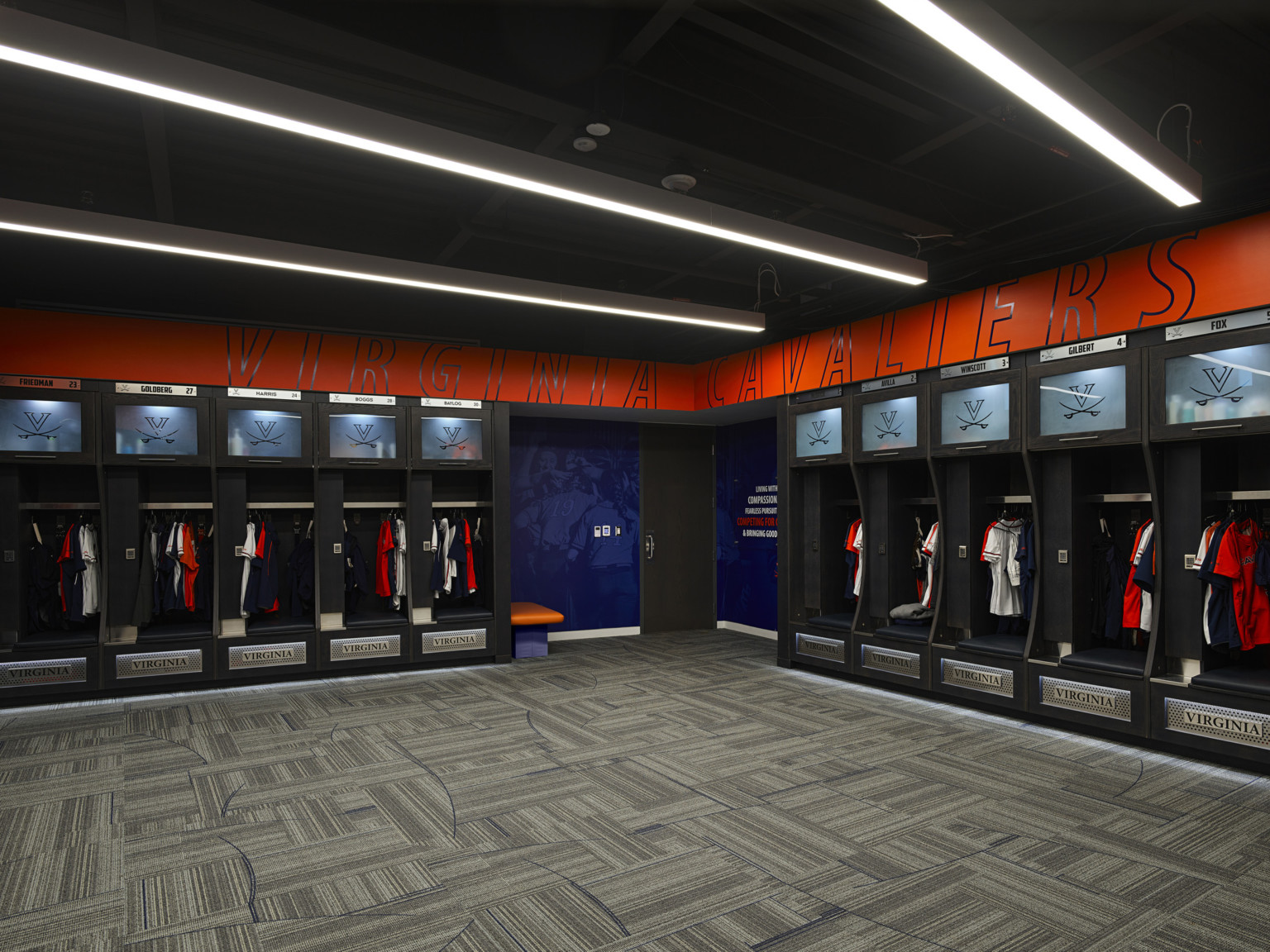 Interior locker room. Virginia Cavaliers written across corner of red wall over open lockers with cushioned seats. Blue mural