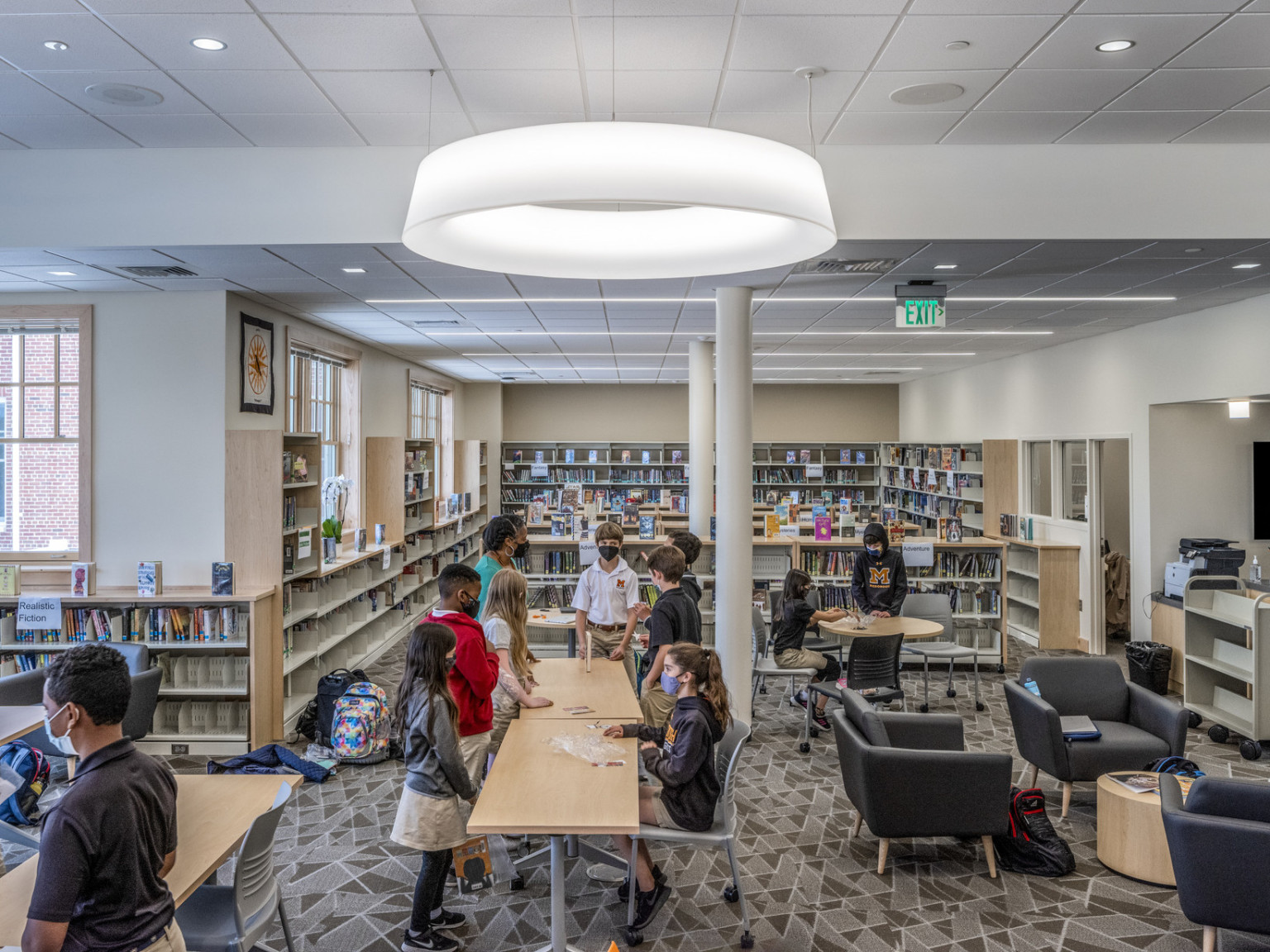 Middle school library with half height bookshelves, flexible tables and seating, arm chairs. Round pendant lights above