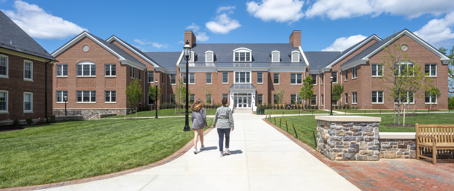 2 students walking down a paved path to white framed front entrance of brick multistory independent school. Grass lawn, seating