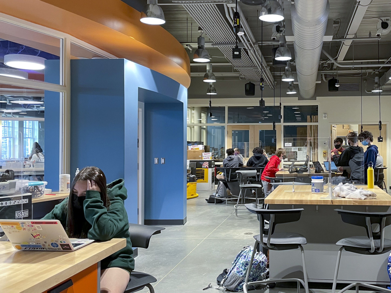 Students working on wood tables in classroom with blue framing at entrance and orange wall trim detail. Exposed ductwork above