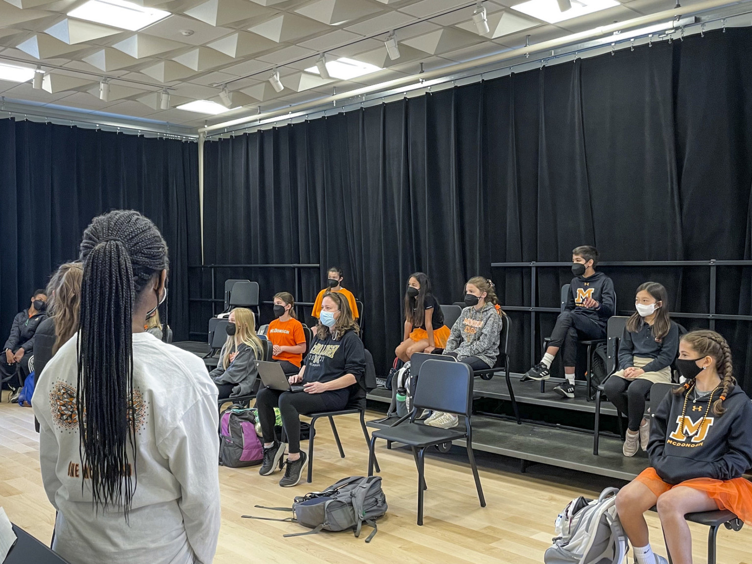 Students in classroom with chairs on risers and black curtains hanging along walls. Geometric acoustic panels on ceiling