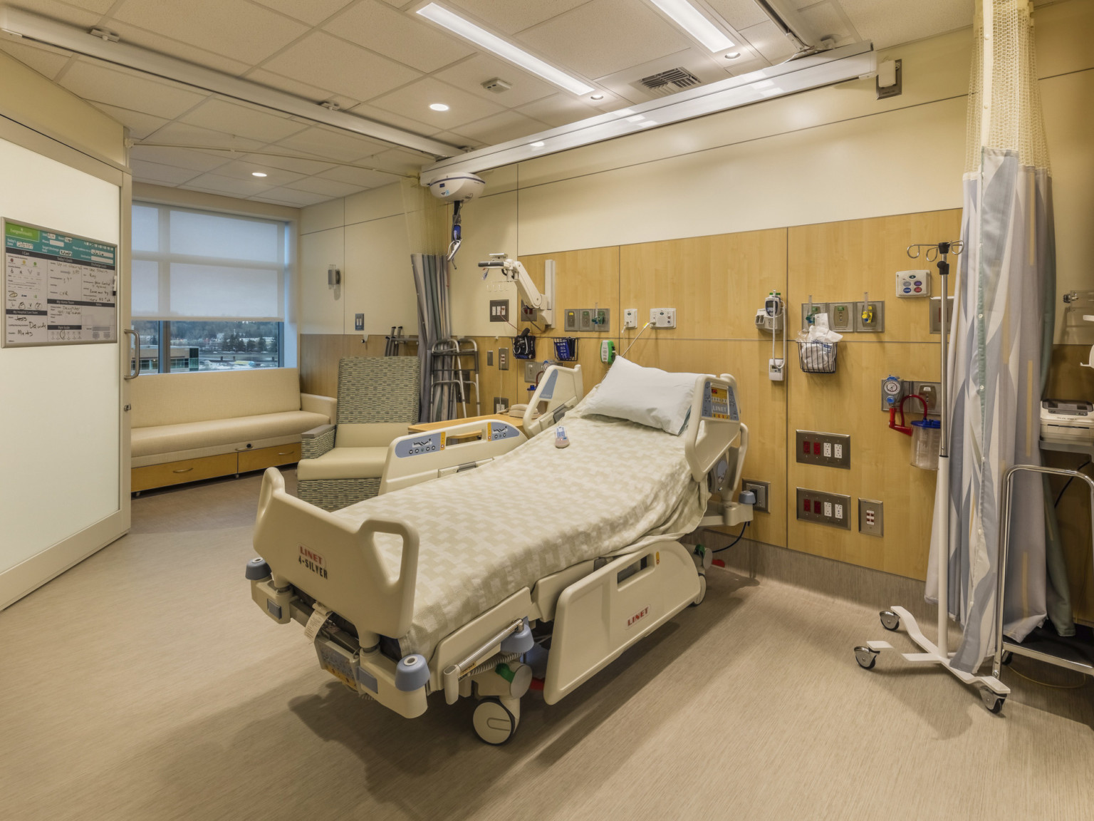 Hospital bed in white room against wall with wood panel accents and medical equipment. Flexible wall curtain at right