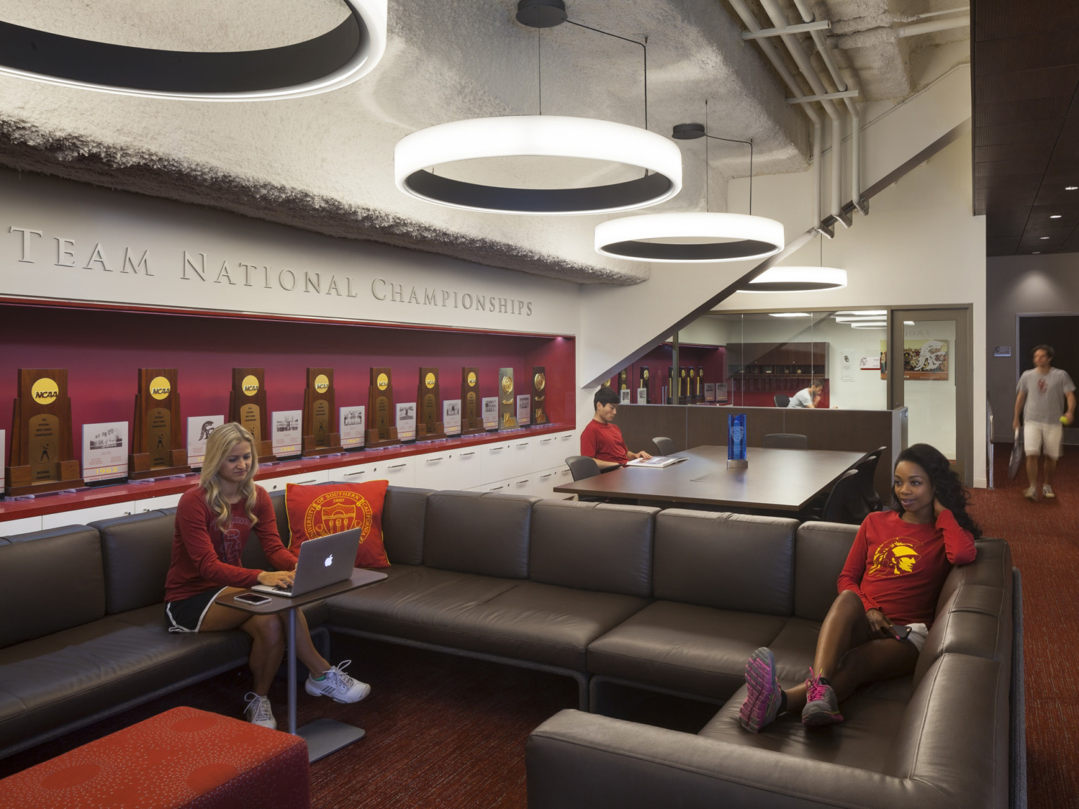 Textural stepped roof with round modern chandeliers over couches and flexible workspaces. Red trophy case to left