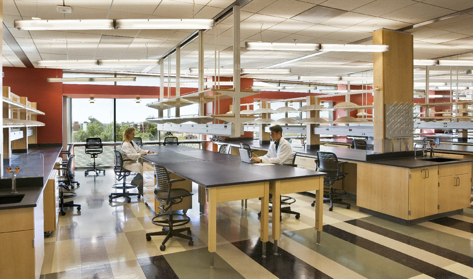 2 people in lab coats at table in laboratory, large windows on back wall, red accent wall, shelves hang over rows of tables