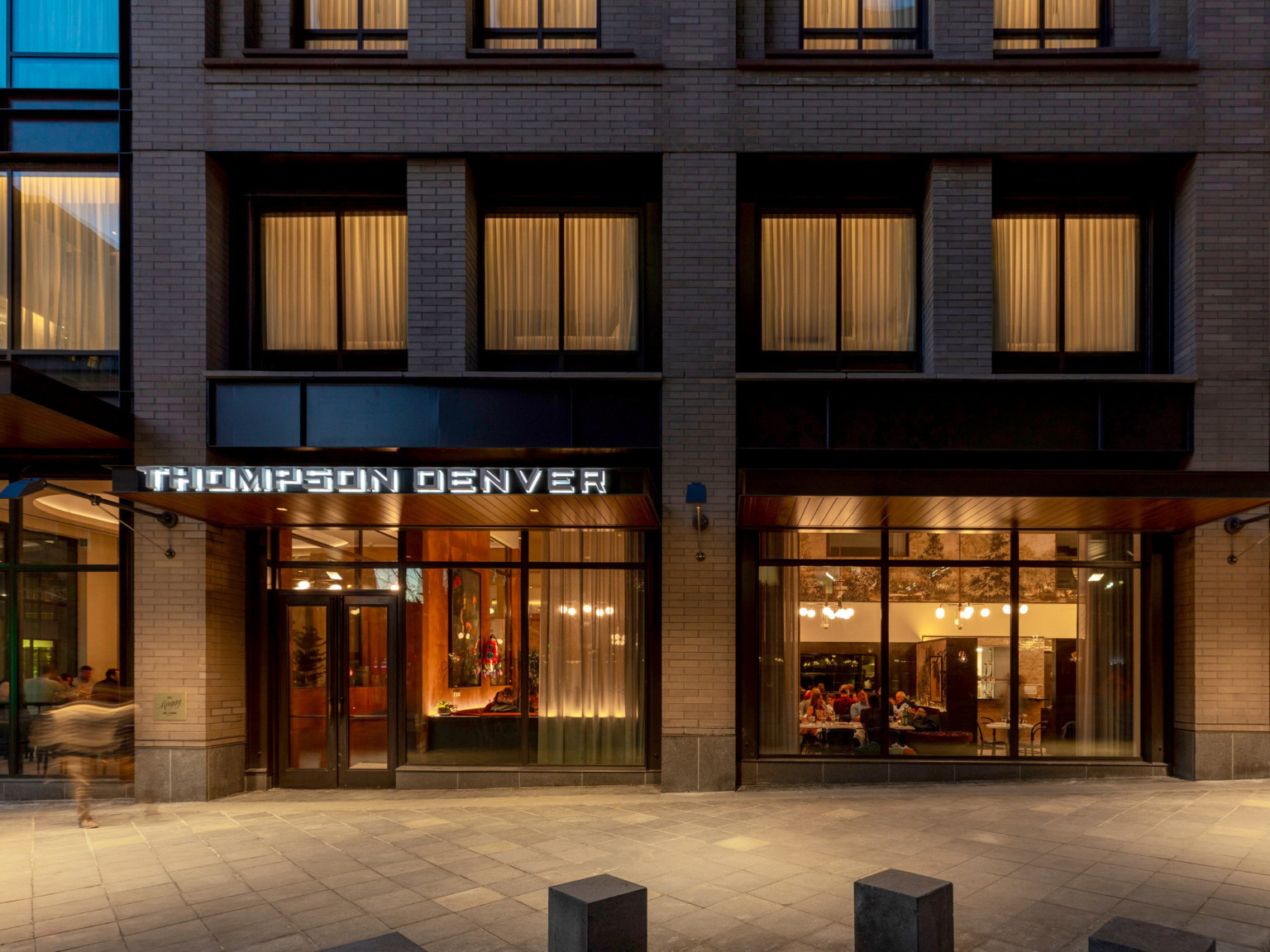 exterior view of entry with floor to ceiling windows, backlit Thompson Denver sign over double doors framed by brick columns