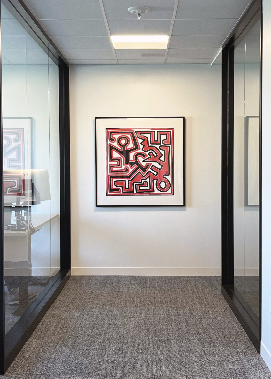 square framed red white and black keith haring print hanging at the end of hallway on white wall between glass movable walls