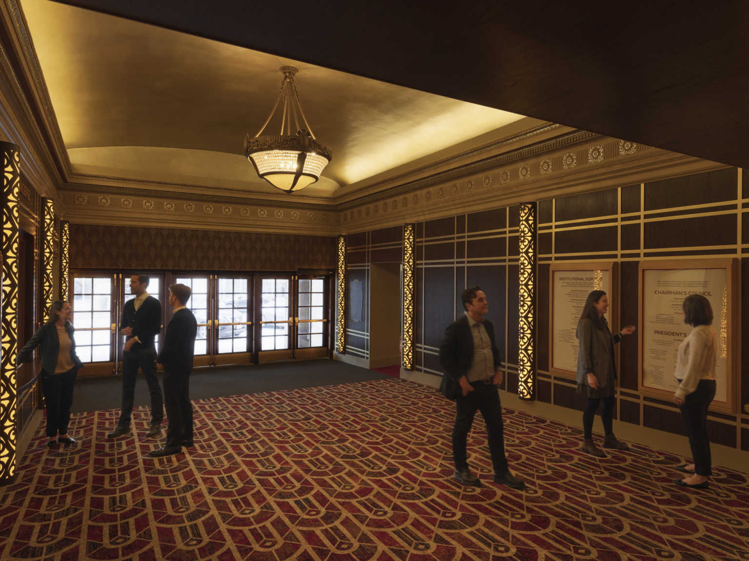 Entry to theater with multiple glass double doors, patterned carpet, information on walls framed by illuminated art deco columns