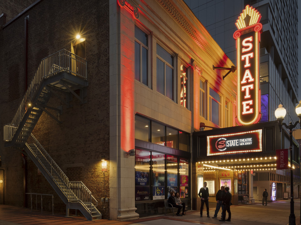 Theater exterior from corner with illuminated retro neon blade sign reading State over marquee. Stairs on side of building