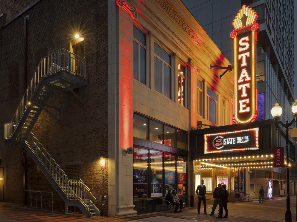 Theater exterior from corner with illuminated retro neon blade sign reading State over marquee. Stairs on side of building