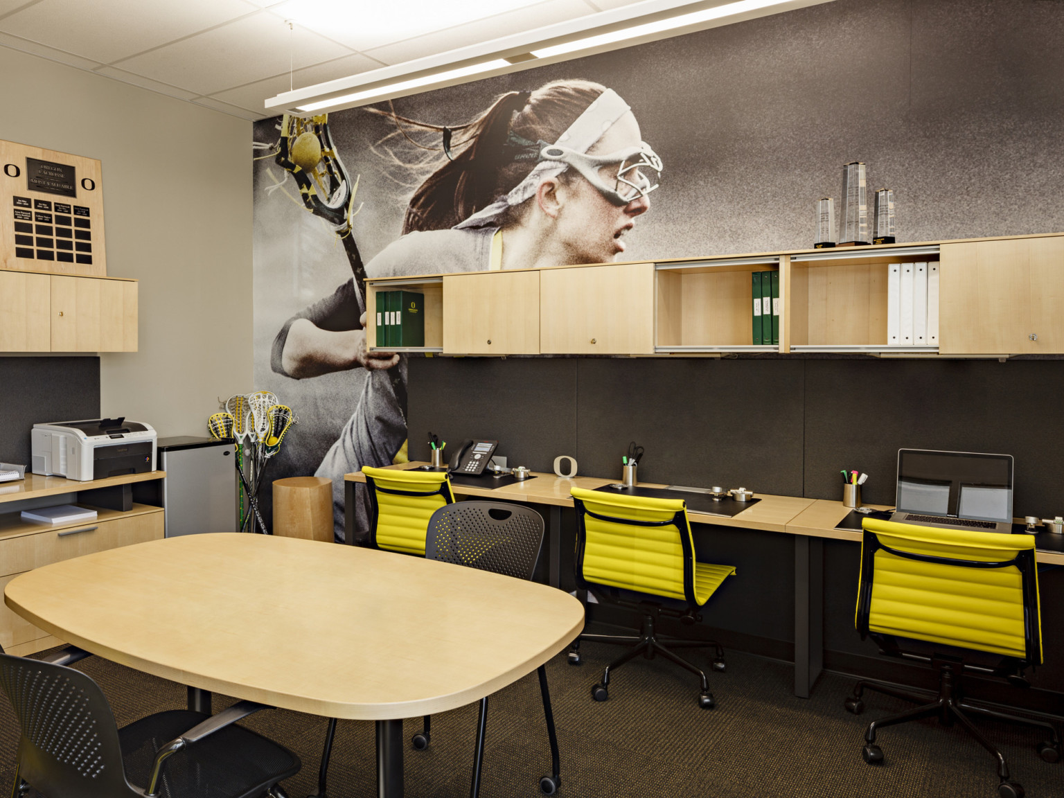 Room with lacrosse player photo mural on right wall. Flexible workstations along both walls, wood counters and cabinets, table