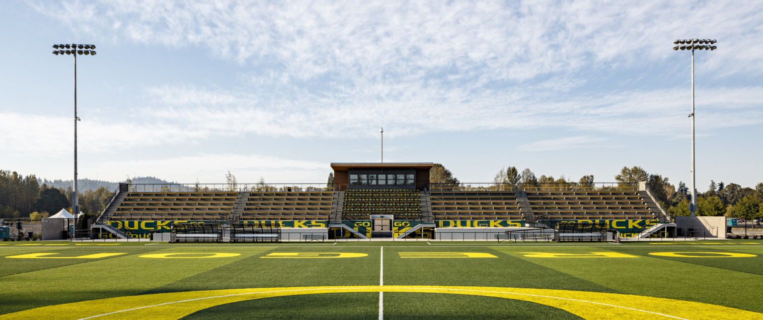 Papé Field seen from center line during game, green and yellow bleachers reading Go Ducks. Press box with canopy extending