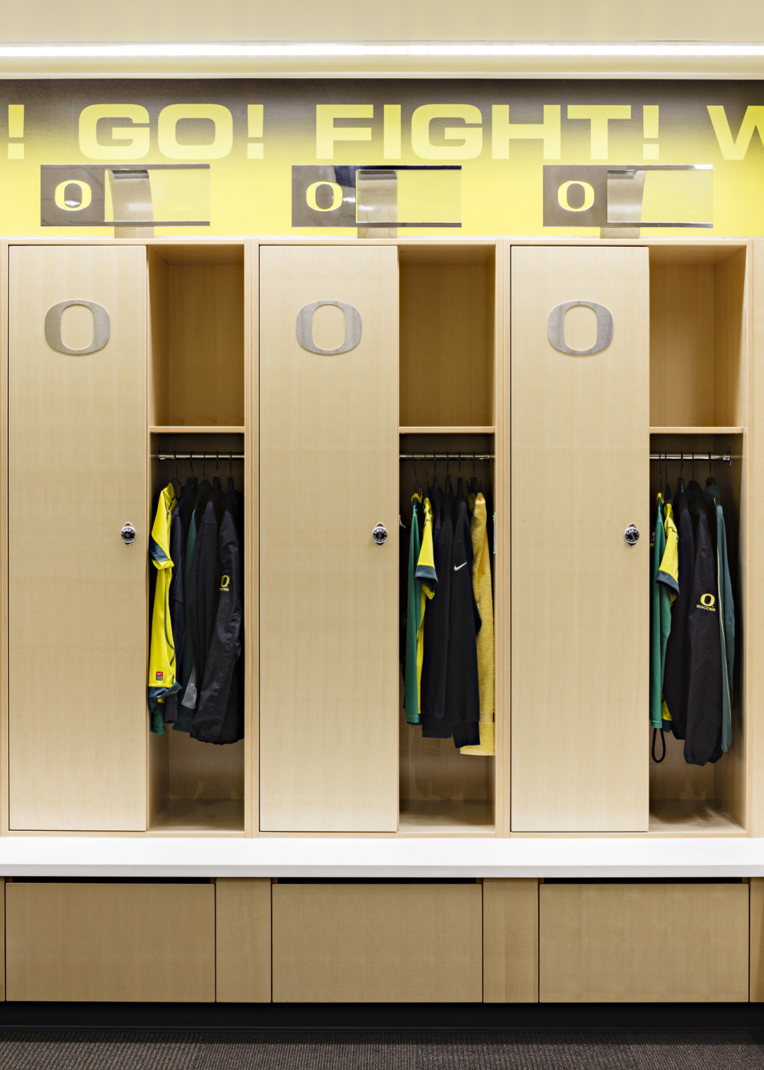 Closeup of wood lockers with Oregon logo on doors and exposed hanger and shelf. White bench. Above, fight song and nameplates