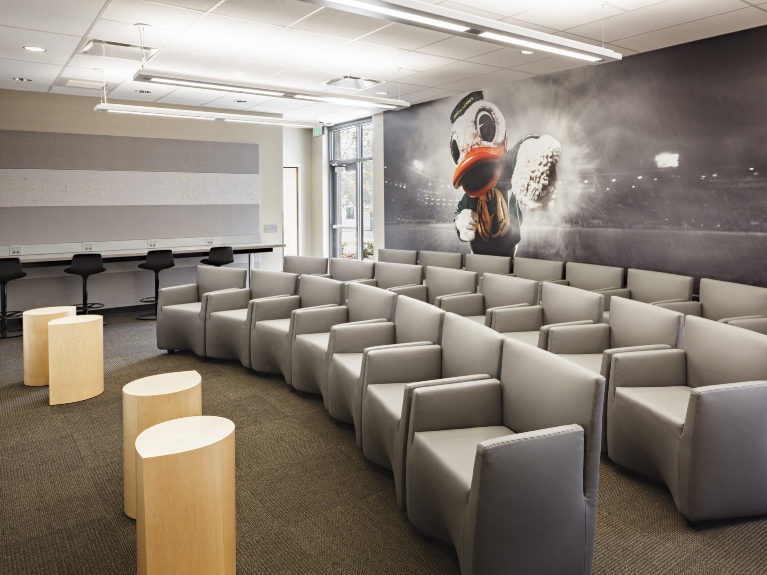Oregon Duck mascot photo mural on wall behind rows of grey armchairs facing small wood tables. Work counter at far wall
