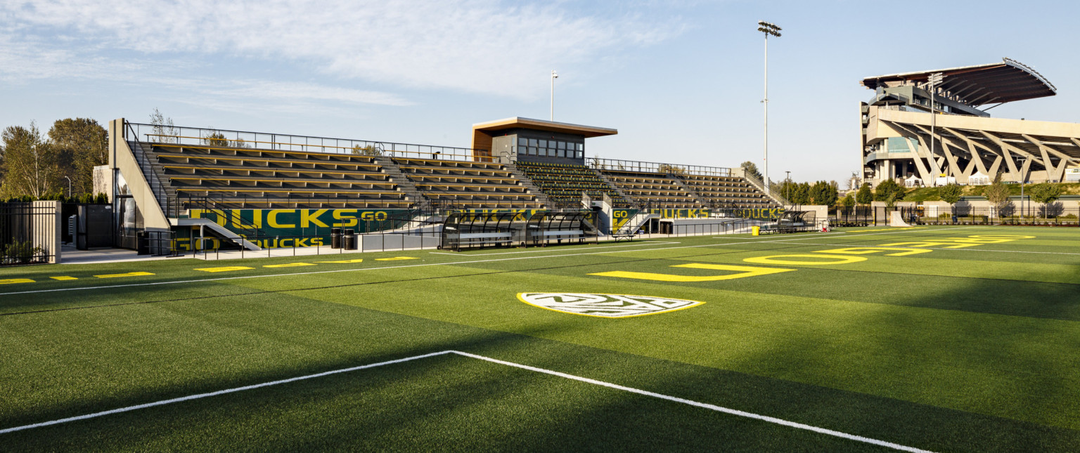 Field seen from goal box looking towards green and yellow bleachers. Brown paneled press box, canopy extending at center line