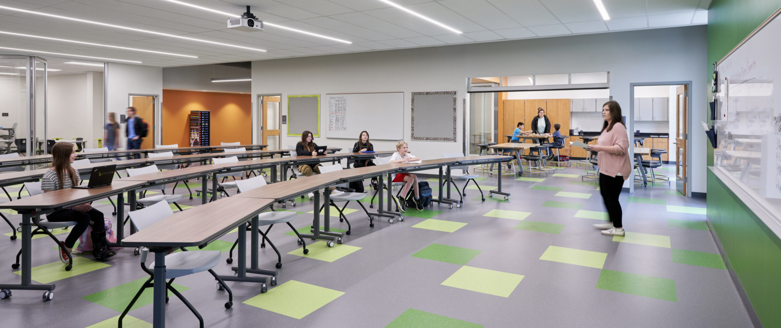 Classroom with green accent wall and floor tiles. Orange walls in hallway through flexible walls. Rolling chairs and long tables