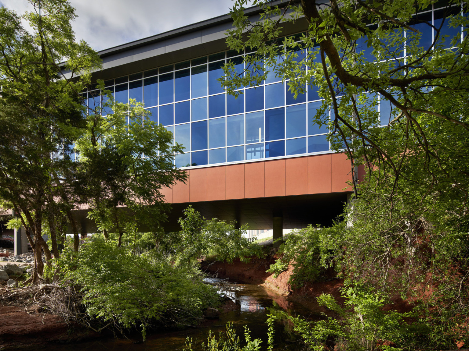 Bridge over stream with double height windows surrounded by trees. Blue tinted glass on orange building