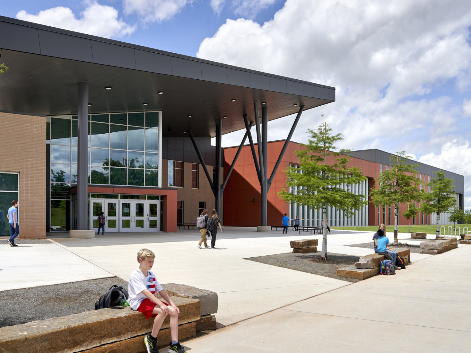 Capps Middle School front entry with angled black shelter over double height glass lobby. Stone benches. Orange wrap facade