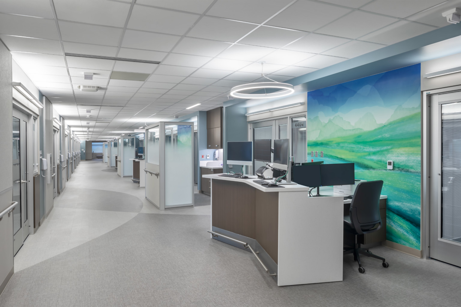 Hospital corridor with angled desk with multiple monitors in front of abstract blue and green mural. Glass partitions beyond