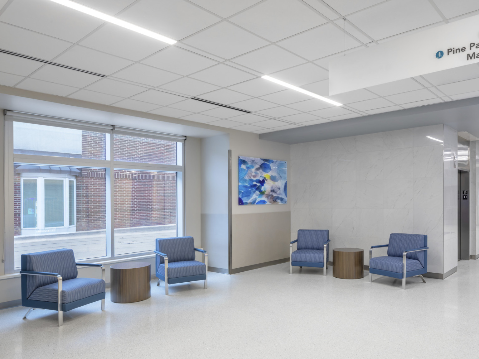 Hospital waiting room with large window looking out to brick wall and bay window. Blue arm chairs in white hallway, elevators