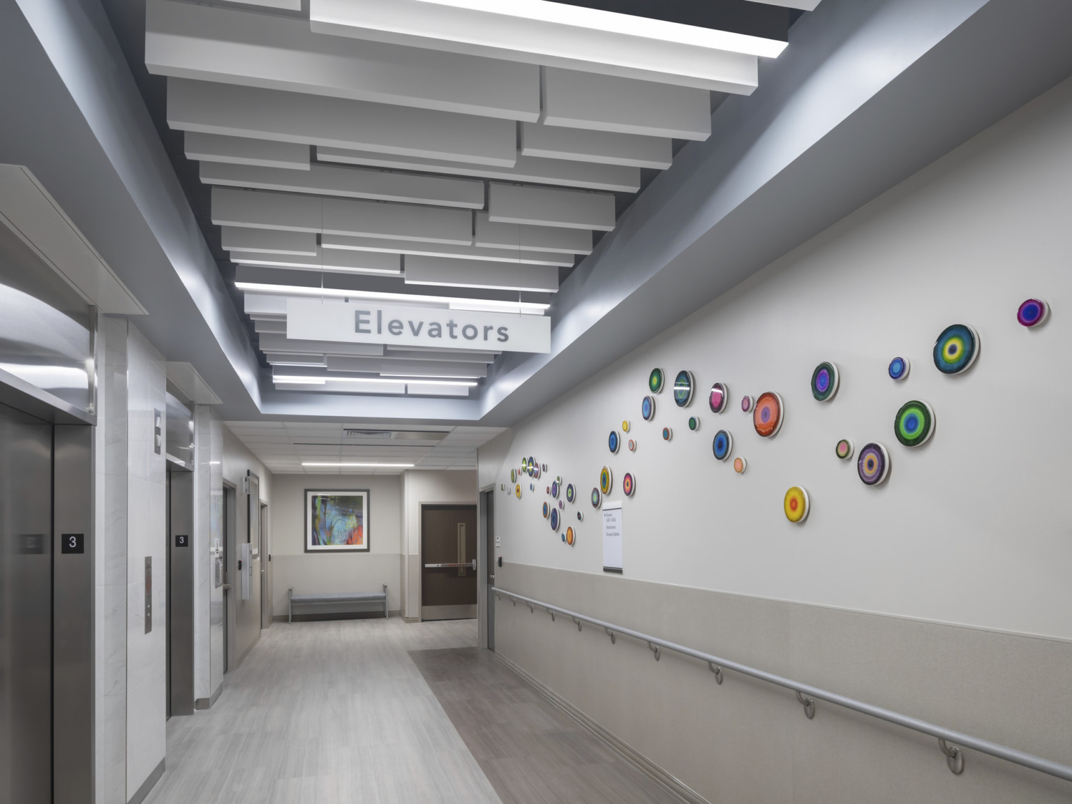 White and grey hallways with acoustical panels hanging above, abstract round art on wall opposite elevators