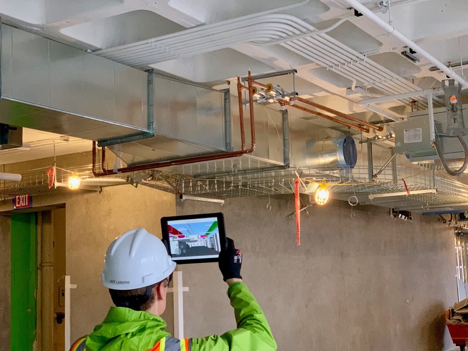 person in bright green safety jacket and construction hat with handheld screen device up at ceiling pipes on construction site