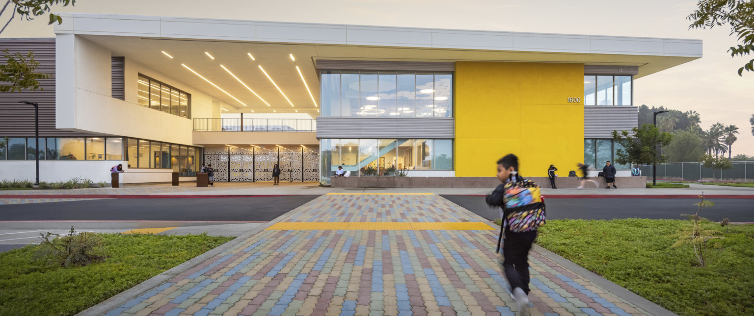 Front entrance of elementary school with multicolor stone path leading to yellow building with grey accents and white screen gates