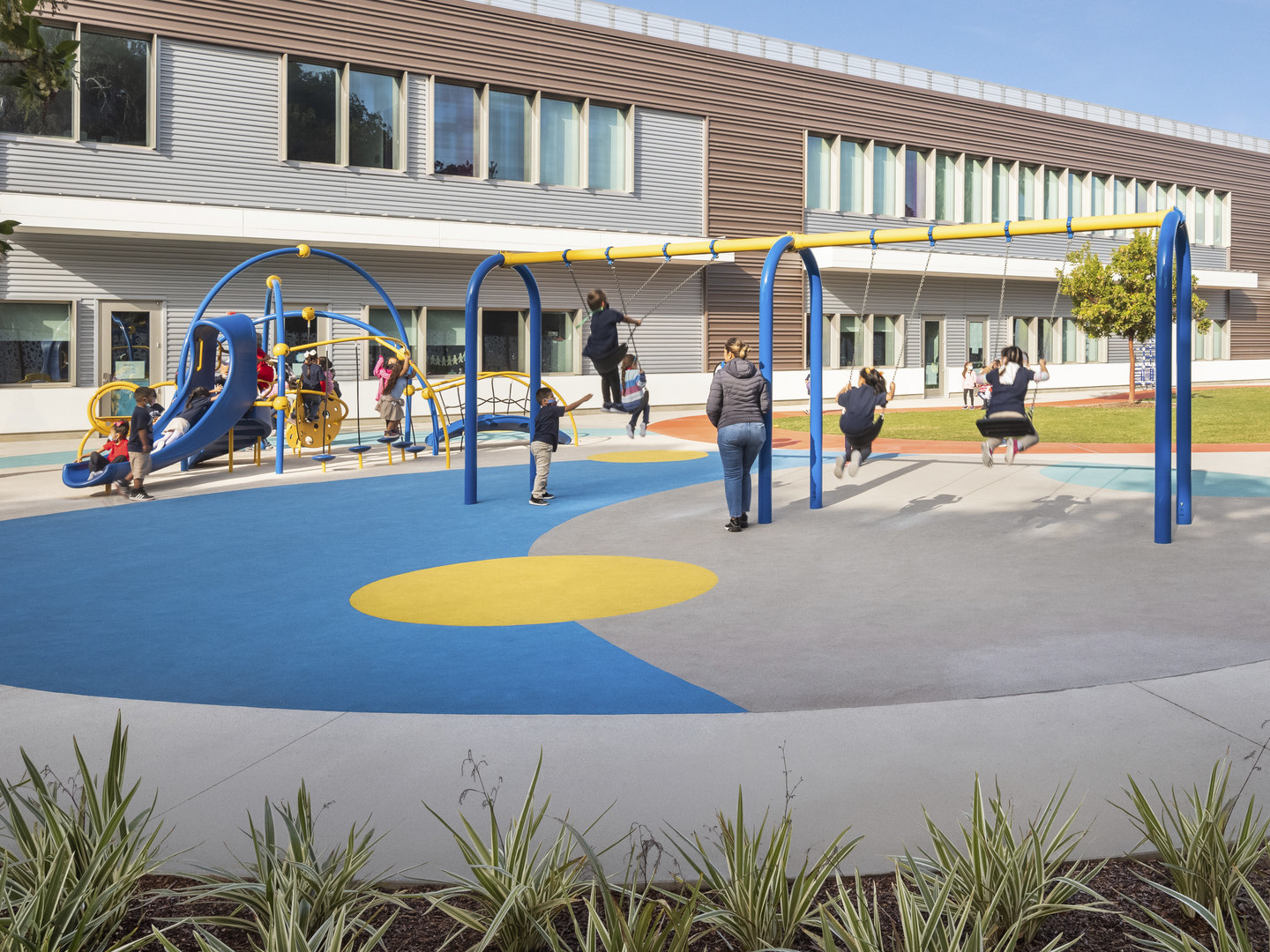 Blue and yellow playground with geometric abstract pattern to padded ground. Surrounded by greenery and backed by 2 story school