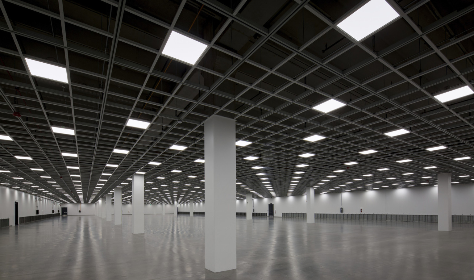 wide open room with reflect cement floors, white square columns white walls, open black ceiling with repeating light squares