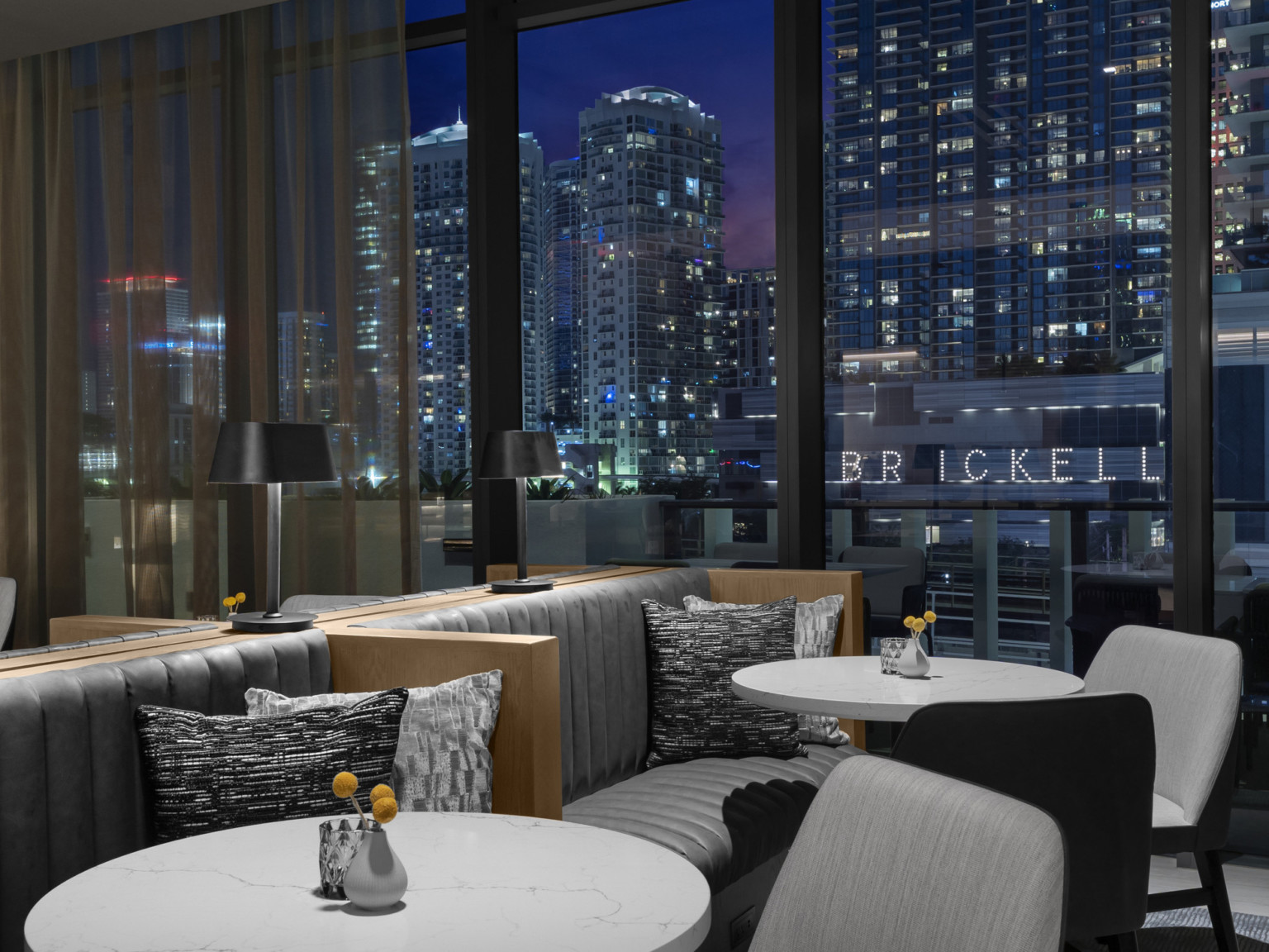 View of the city lights at night through floor to ceiling windows in AC Hotel lounge, mixed booth and chair seating at round white tables