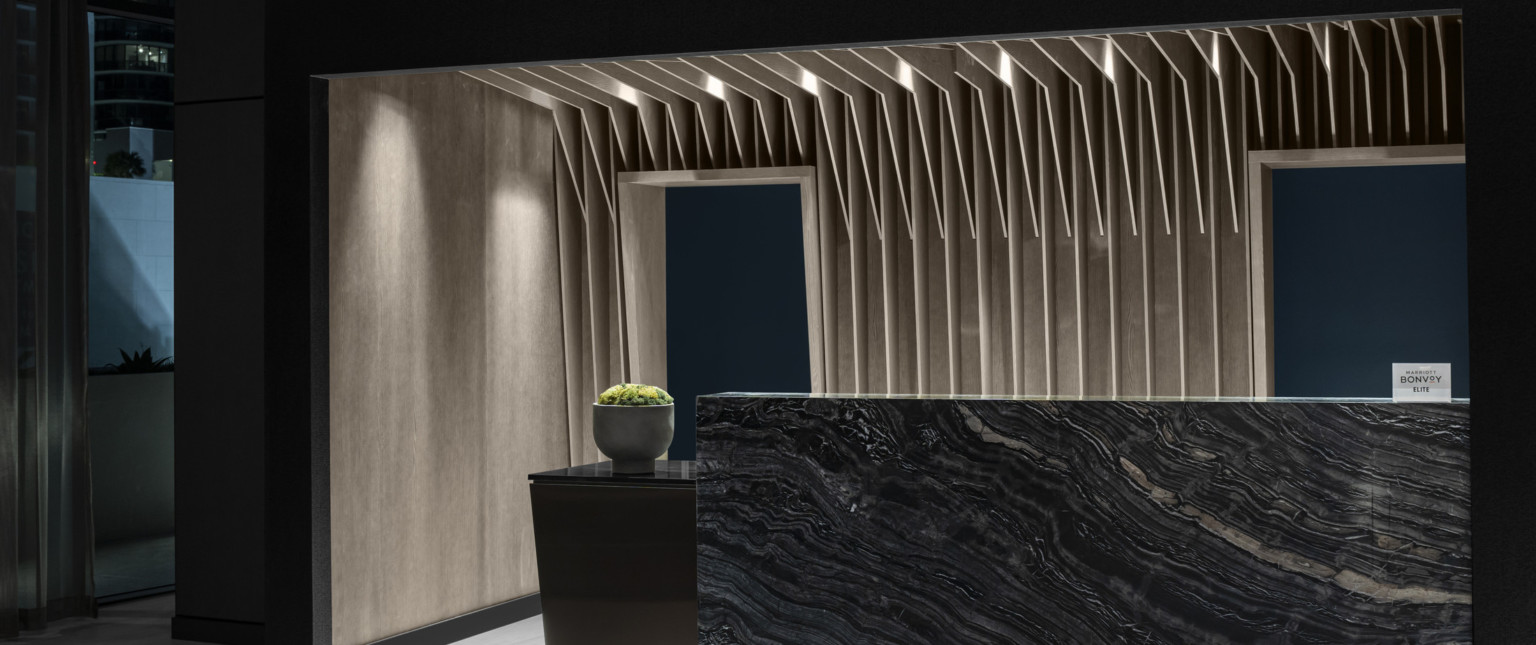 The AC Hotel reception desk, a dark stone slab with warm wood slats like palm fronds vertically emerge overhead in recessed space The AC Hotel reception desk, a dark stone slab with warm wood slats like palm fronds vertically emerge overhead in recessed space