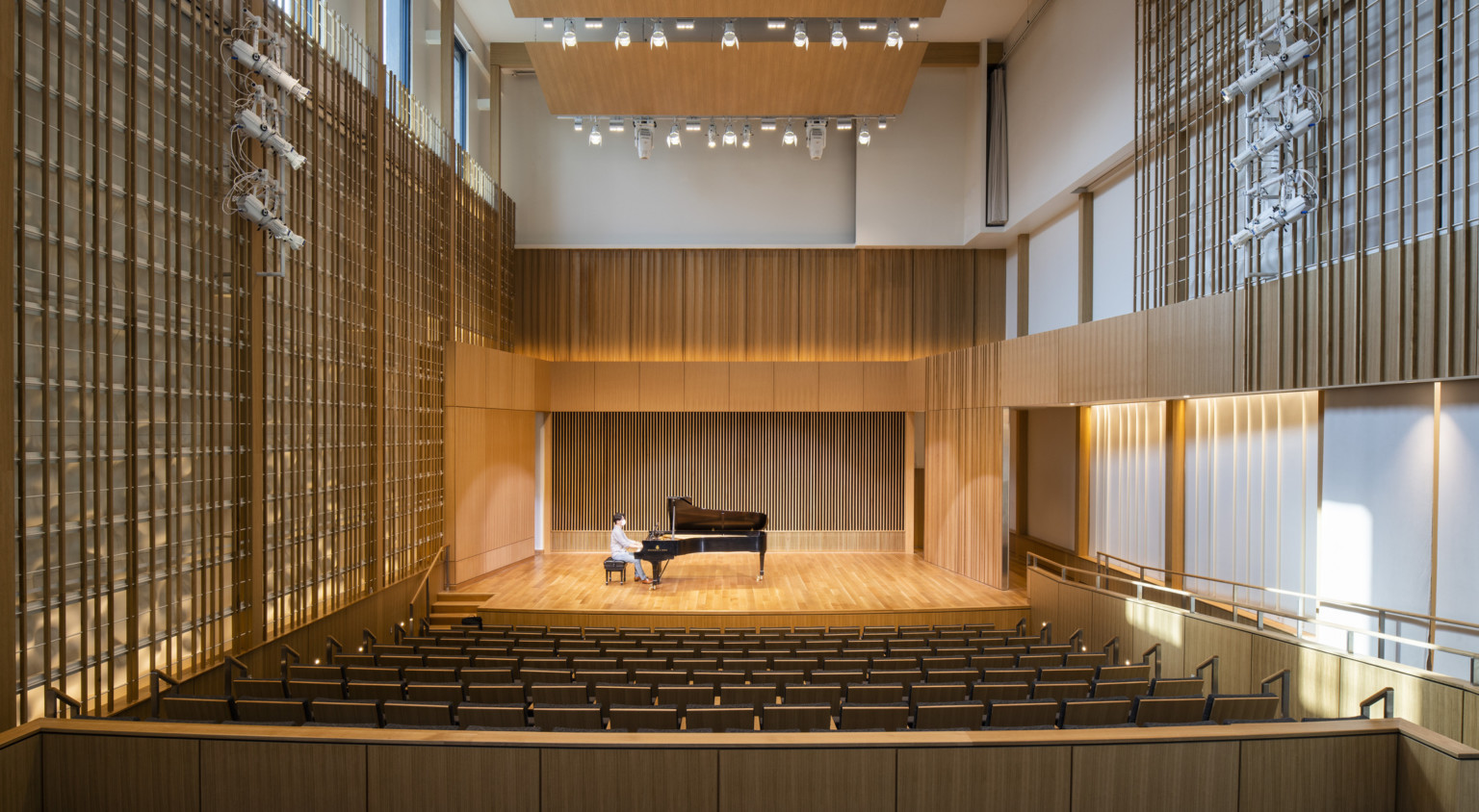 Man sitting at piano in double height recital room auditorium with theater seating. Wood panel designs on walls and wood acoustic panel