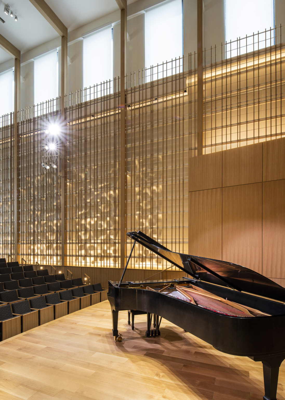 Angled view from side of stage looking toward wood design on wall illuminated from behind. Grey theater seats with light wood
