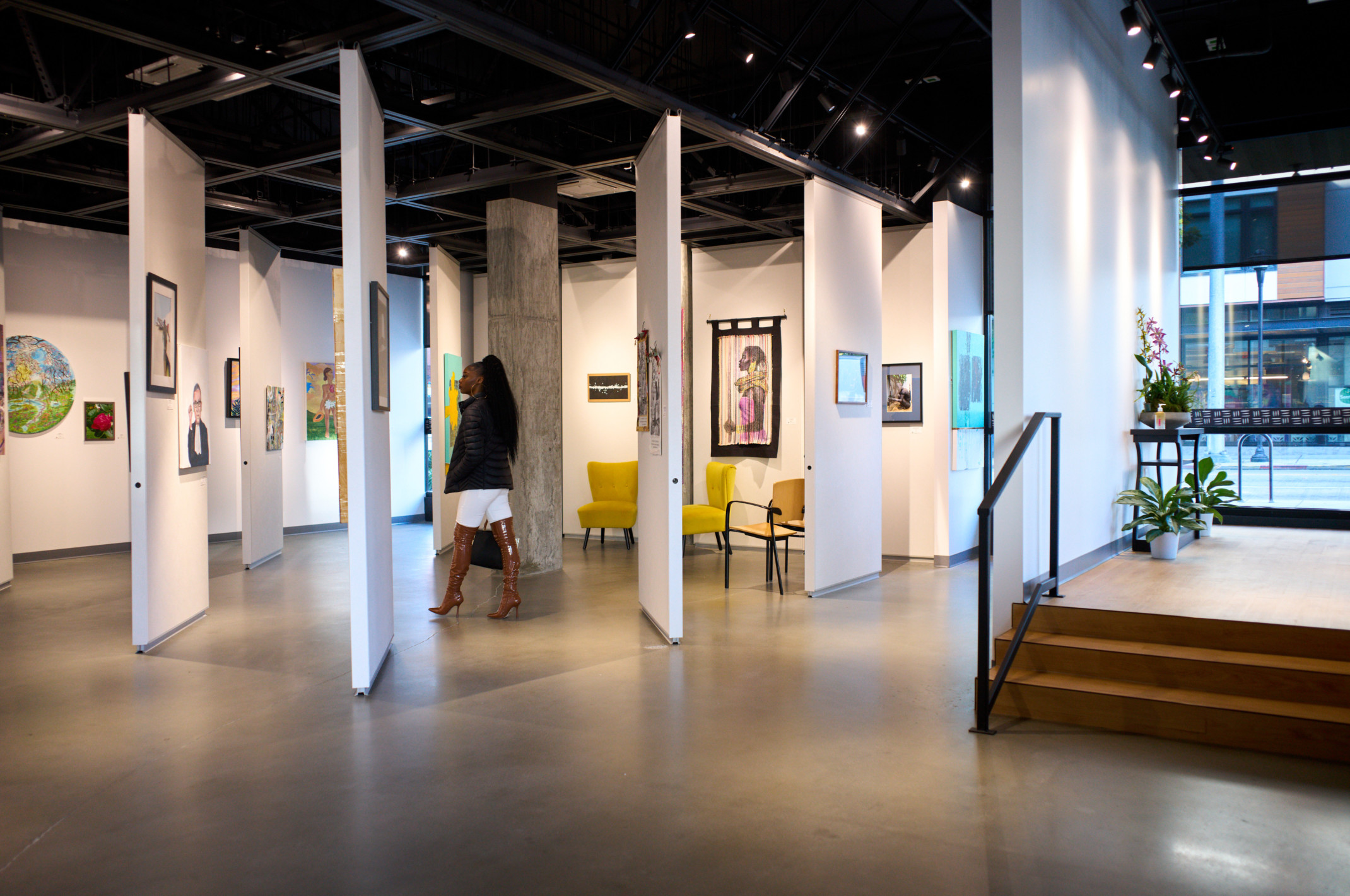 Woman standing in Arte Noir Gallery interior in Seattle, movable white wall panels hang art in room with floor to ceiling windows, black ceiling Woman standing in Arte Noir Gallery interior in Seattle, movable white wall panels hang art in room with floor to ceiling windows, black ceiling